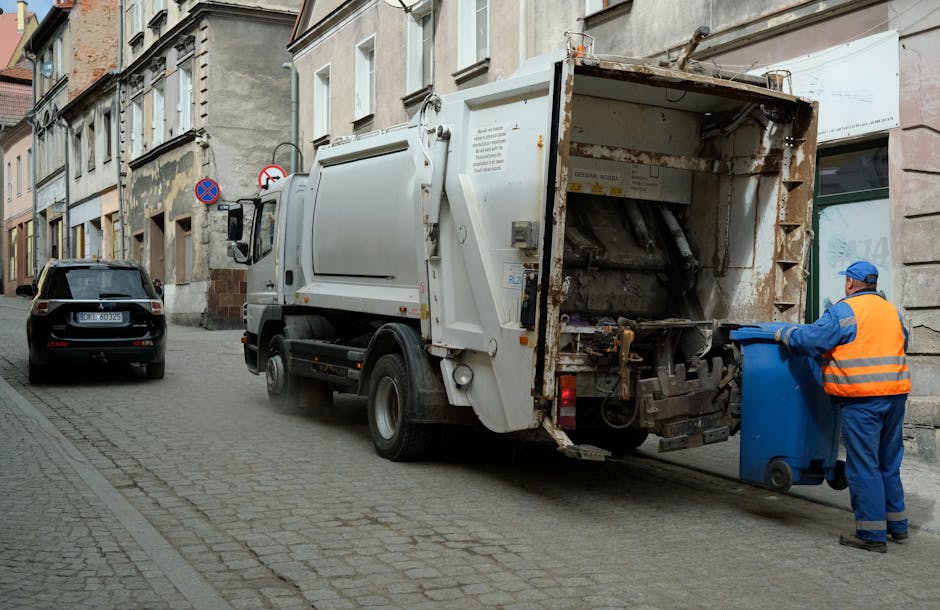 A waste collection vehicle, painted white with some visible rust and dirt on its rear compartment, is positioned on a narrow cobblestone street next to old, multi-story buildings with weathered facades. The truck's rear open loading area reveals mechanical components and rusted metal surfaces designed for compacting household or commercial rubbish. To the right of the vehicle, a worker dressed in a blue uniform and high-visibility orange vest, along with a matching blue cap, is actively emptying a blue wheeled rubbish bin into the truck, ensuring efficient waste disposal. In the background, a black SUV is parked on the street, and above, there are street signs and windows with white frames, indicating an urban environment. The scene is illuminated by natural daylight, emphasizing the textures of the cobblestone street, the rusted metal parts, and the contrasting vibrant safety gear of the worker. This image depicts an independent waste collection activity, characteristic of private rubbish removal services operating in residential neighborhoods consistent with rubbish clearance practices outside of local authority recycling systems.