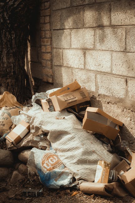A pile of mixed waste materials situated at the base of a brick and concrete wall, including flattened cardboard boxes with visible corrugated texture, partially torn and creased, alongside crumpled brown paper bags and plastic packaging. The waste is gathered on a rough, uneven surface with small stones and dirt, and some bags appear to be resting on or partially covered by a large, light-colored bag or textile material. To the left, a tree trunk is partially visible, suggesting the scene is outdoors, possibly on a pavement or alleyway. The lighting is natural, casting soft shadows, and the arrangement indicates a collection of refuse awaiting removal, which could be sorted or handled through alternative waste disposal methods by a private rubbish clearance service such as Rubbish Clearance Hampstead. The scene visually emphasizes the need for onsite waste collection or removal from private premises, supporting the context of independent rubbish clearance.