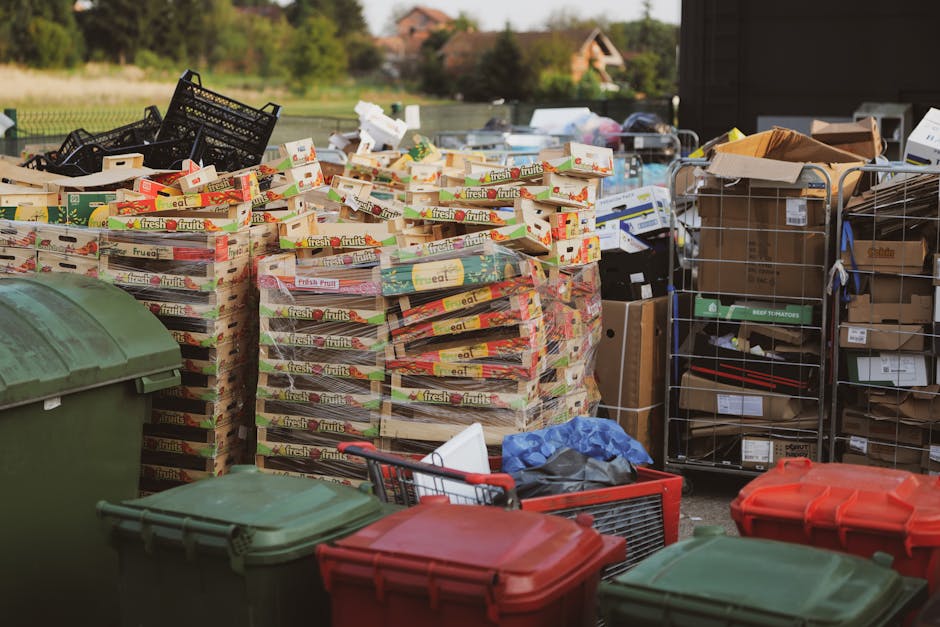 A large outdoor area shows multiple stacks of flattened cardboard boxes labeled with 'fresh fruits,' neatly arranged on a paved surface, with some boxes slightly leaning. Surrounding the stacks are several plastic waste bins in green and red with closed lids, positioned in front and to the sides of the cardboard, indicating waste separation. To the right, a metal trolley filled with various boxes and packaging materials, some missing labels, and a brown paper bag on top, is visible. In the background, additional cardboard boxes and packaging materials are piled on metal racks, along with a blue tarp partially covering items. The setting appears to be an outdoor waste collection or rubbish clearance operation, with natural daylight illuminating the scene, and a hint of greenery and residential buildings in the distance, consistent with private waste collection services such as those offered by Rubbish Clearance Hampstead, supporting alternative rubbish removal options in NW3 areas.