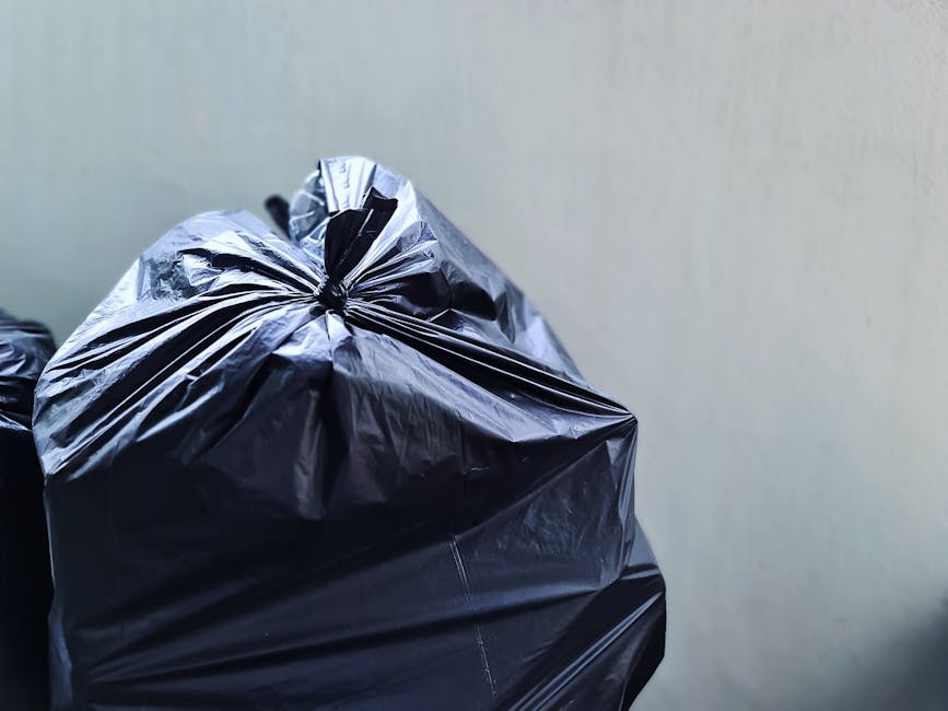 A close-up view of a tightly tied black plastic rubbish bag with a glossy finish, placed against a plain, light-colored wall. The bag appears to be filled with waste material, causing it to bulge slightly and creating creases and folds on its surface. The top of the bag is gathered and sealed with a knot, indicating it has been prepared for collection or disposal. Part of another similar black bag is visible on the left side of the frame. The image is well-lit, highlighting the reflective quality of the plastic material and the texture of the bag's surface. This scene suggests a scenario of private or independent waste handling, aligning with rubbish removal services provided by companies such as Rubbish Clearance Hampstead, particularly for local or on-site rubbish clearance, as often required for home or small business waste management.