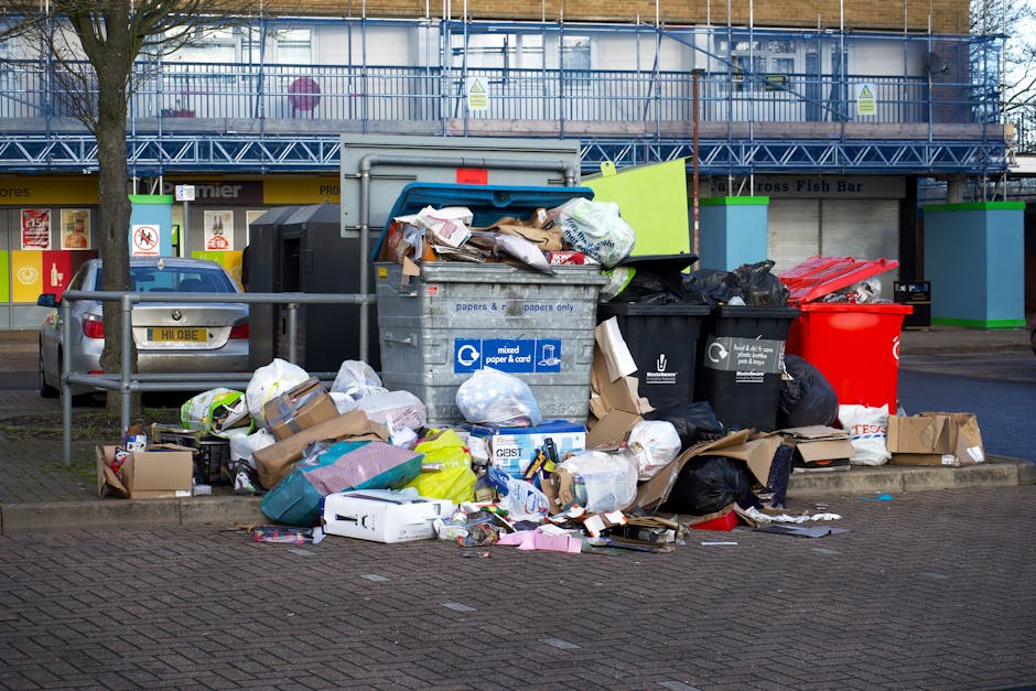 A large outdoor area shows multiple stacks of flattened cardboard boxes labeled with 'fresh fruits,' neatly arranged on a paved surface, with some boxes slightly leaning. Surrounding the stacks are several plastic waste bins in green and red with closed lids, positioned in front and to the sides of the cardboard, indicating waste separation. To the right, a metal trolley filled with various boxes and packaging materials, some missing labels, and a brown paper bag on top, is visible. In the background, additional cardboard boxes and packaging materials are piled on metal racks, along with a blue tarp partially covering items. The setting appears to be an outdoor waste collection or rubbish clearance operation, with natural daylight illuminating the scene, and a hint of greenery and residential buildings in the distance, consistent with private waste collection services such as those offered by Rubbish Clearance Hampstead, supporting alternative rubbish removal options in NW3 areas.