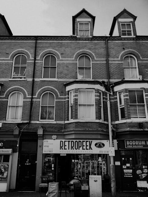 The image depicts a multi-storey brick building with a retail shop on the ground floor, identified by a sign reading 'RETROPEEK HOUSE CLEARANCES' indicating an independent waste clearance service. The building features four large, arched windows on the upper floors, with the top floor having two dormer windows set into the pitched roof, which is covered in dark roofing material. The brickwork is detailed with contrasting lighter mortar joints, and the facade shows horizontal decorative brick bands. In front of the shop, there are several small waste bins and a white postalbox, suggesting collection or disposal points related to the business. A neighbouring shop sign reading 'BODRUM' is partially visible to the right. The scene is captured in black and white, with natural daylight illuminating the building, highlighting textures of the brick and window frames. This setting illustrates a typical high street frontage where private waste clearance services are offered, aligning with the focus on rubbish removal or alternative waste handling methods outside local authority collection services, which is relevant to the Hampstead High Street rubbish clearance guide context.