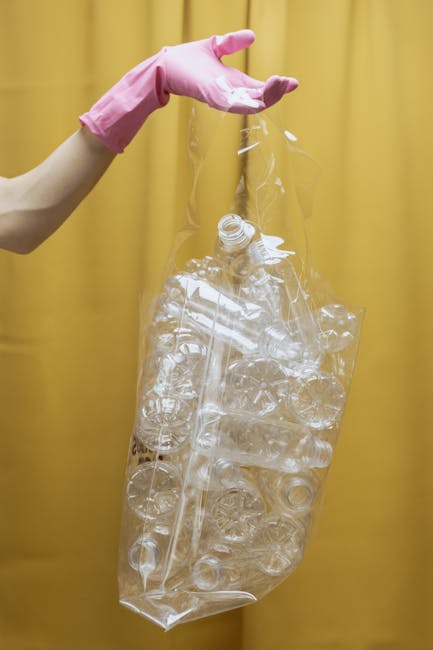 A person's hand wearing a pink rubber glove is holding a clear plastic bag filled with empty plastic bottles and containers, which are crumpled and discarded. The background features a plain yellow curtain or wall, providing a neutral backdrop that highlights the transparent waste materials. The scene emphasizes the process of managing recyclable waste, with the plastic items appearing ready for collection or recycling in an environment where waste separation and private disposal services, such as those offered by Rubbish Clearance Hampstead, may be involved. The focus is on the disposal of lightweight, destructured plastic packaging, reflecting standard practices in waste sorting and environmentally conscious rubbish removal. The lighting is even, with no harsh shadows, giving a clear view of the plastic debris's textures and transparency, reinforcing the context of rubbish handling and collection for recycling or disposal.