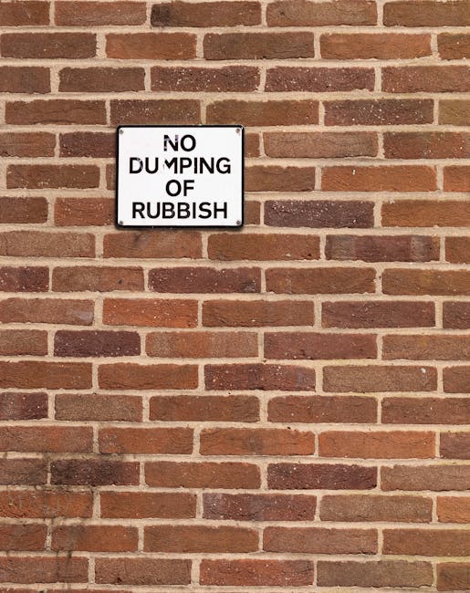 A rectangular metal sign with a white background and black text reading 'NO DUMPING OF RUBBISH' is affixed to a red brick wall. The brick wall has a traditional running bond pattern with reddish-brown and dark brown bricks interspersed, and visible mortar joints. The sign is positioned slightly above the middle of the image and appears to be securely mounted. The environment suggests an exterior setting, possibly in an urban or residential area where rubbish clearance or waste disposal regulation is relevant. The lighting is natural, casting subtle shadows on the wall that highlight the texture of the bricks, which have a slightly weathered finish. There are no other objects or markings visible in the image, emphasizing the signage’s role in discouraging improper rubbish disposal and reflecting an environment where private waste management solutions, such as independent rubbish removal services, may be employed, aligning with the context of alternative waste handling options provided by companies like Rubbish Clearance Hampstead.